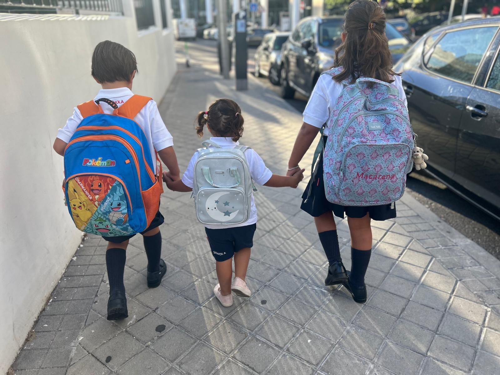 Tres niños con uniforme y mochilas escolares caminan de la mano de espaldas hacia el colegio