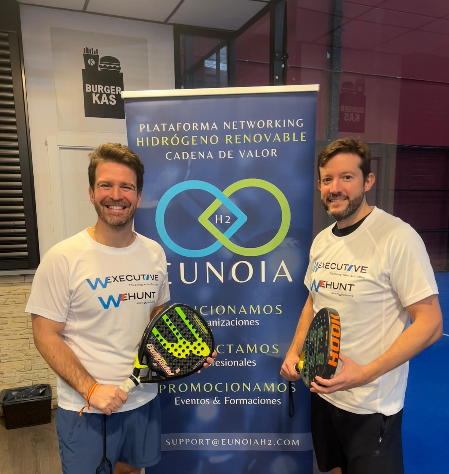 Dos hombres con palas de padel y camisetas W Executive y WeHunt ante un roll-up de Eunoia H2