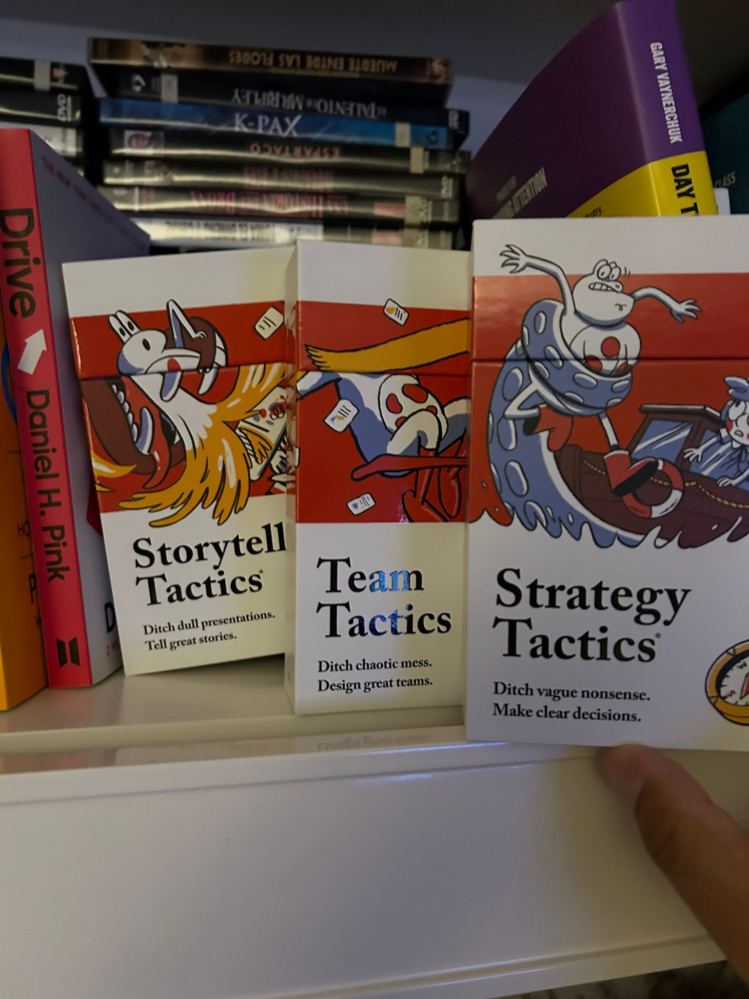 Cajas de las barajas Storytell Tactics, Team Tactics y Strategy Tactics junto al libro Drive en una estantería