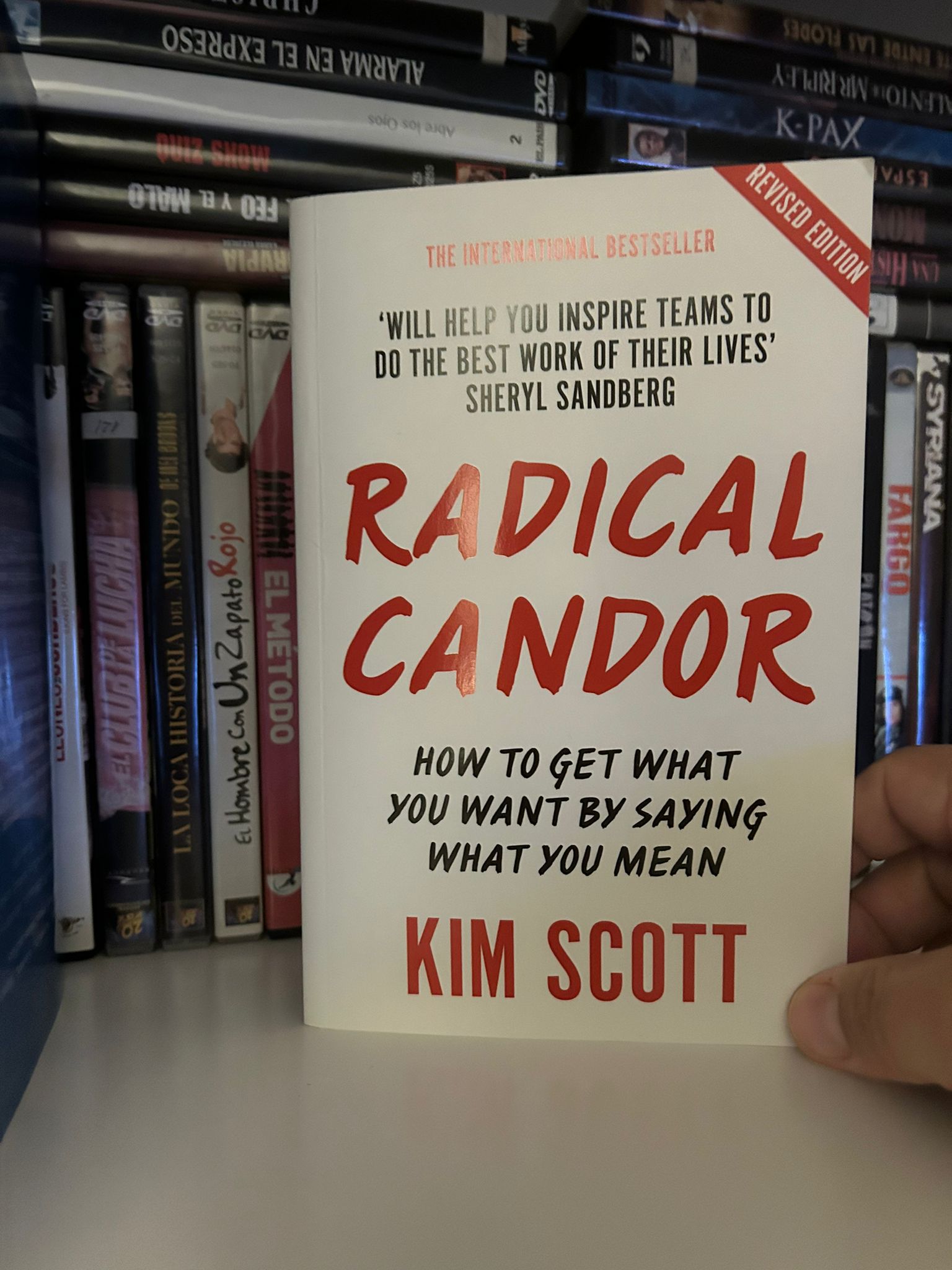 Ejemplar del libro Radical Candor de Kim Scott sostenido frente a una estantería de DVDs