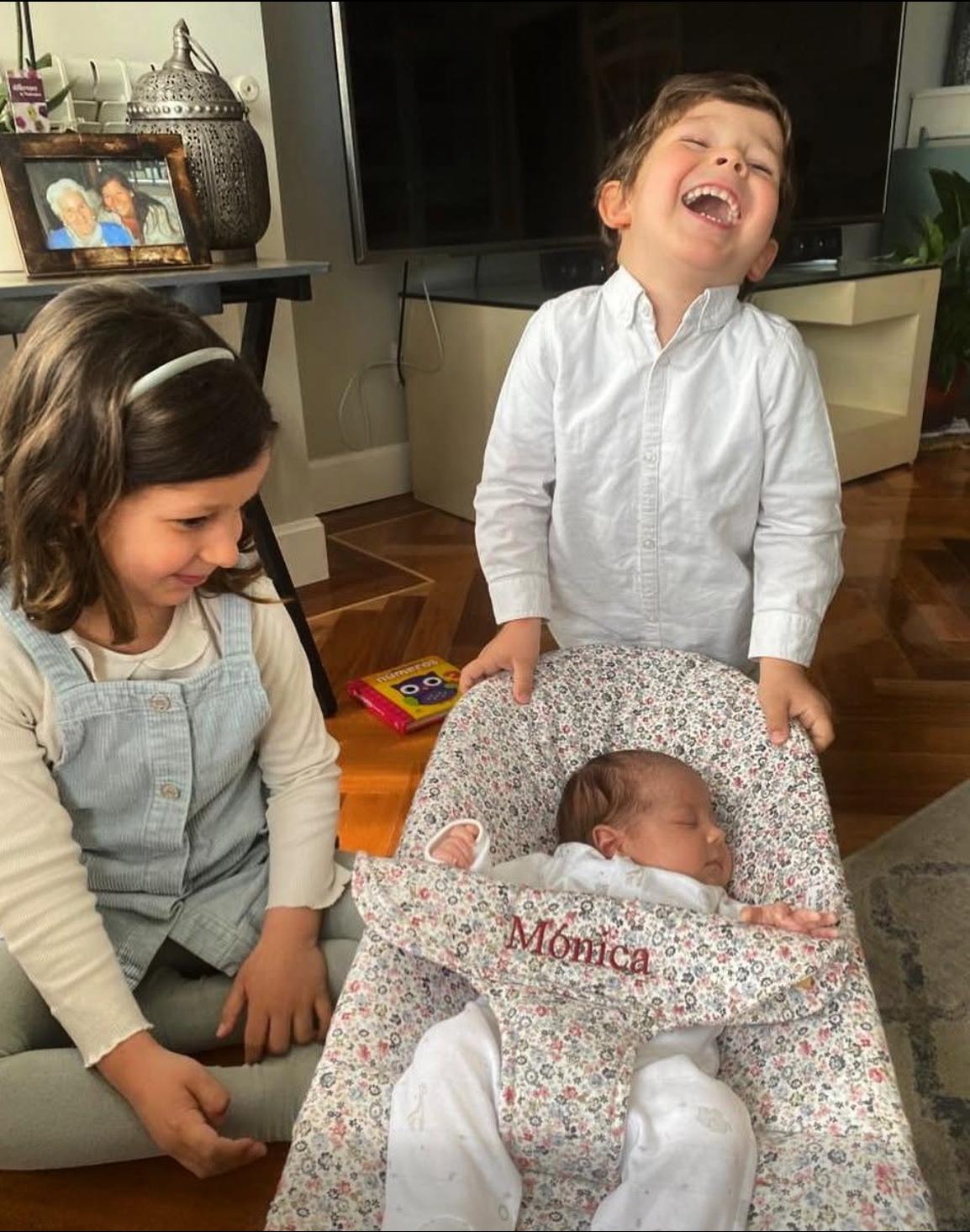 Dos niños sonrientes junto a su hermana recién nacida en un capazo con el nombre Mónica