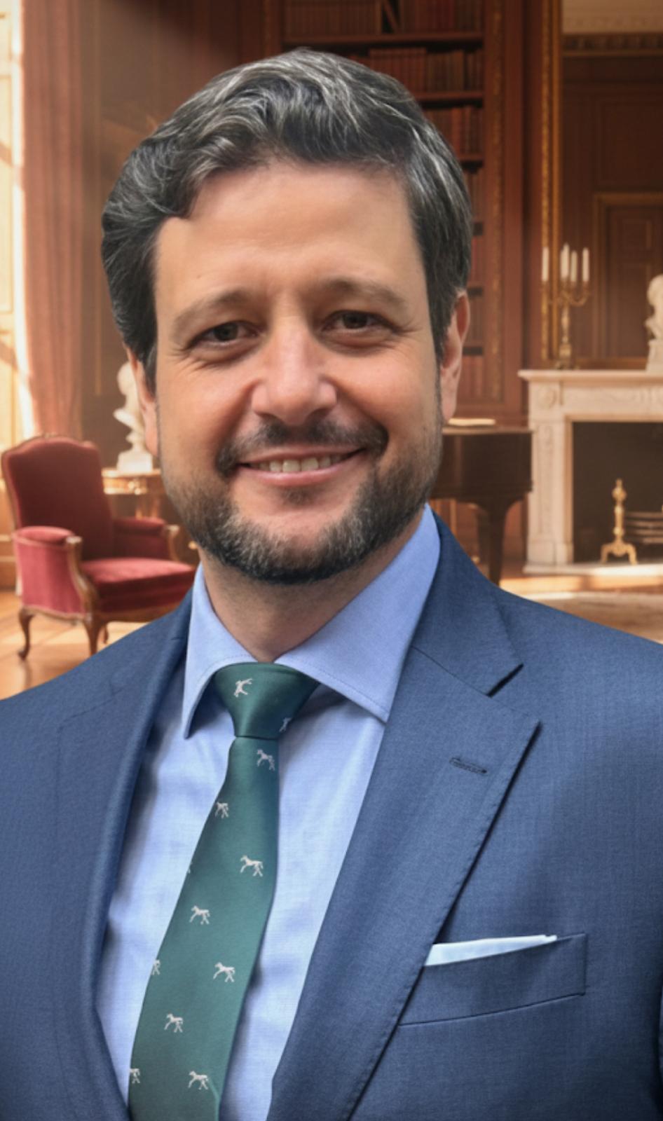 Retrato de Manu Soriano con traje azul y corbata verde en una biblioteca clásica
