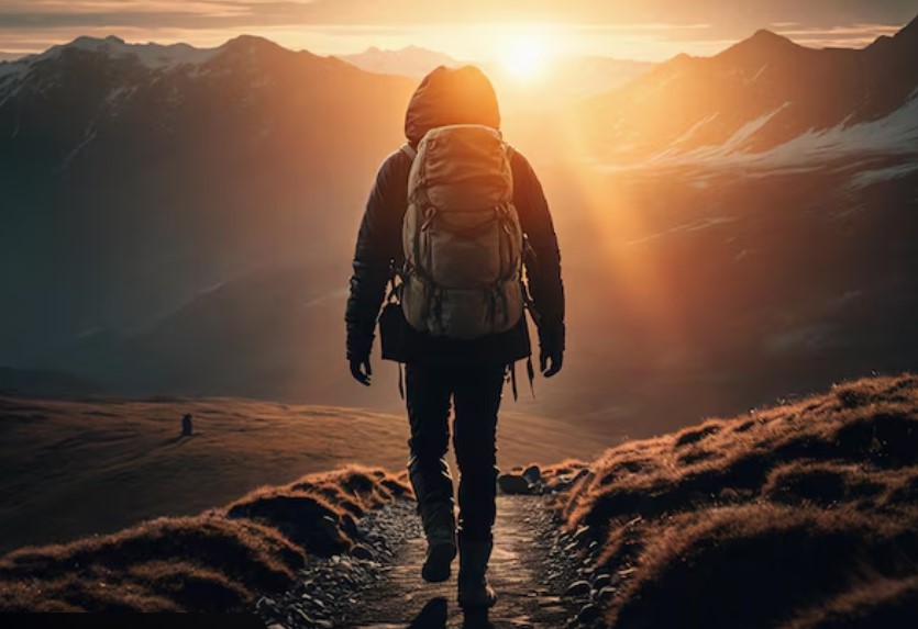Excursionista de espaldas con mochila caminando hacia el sol naciente entre montanas
